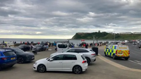 Carla Fowler/BBC Cars parked on the prom at Scarborough with lots of people around and a police van on the road 