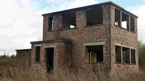 A former RAF watchtower is surrounded by overgrown shrubs. It has no windows and is two storeys high with red and brown bricks.