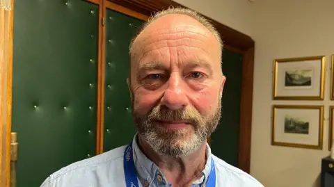Bill Nigh has receding, greying hair and a short beard. He wears a lanyard over a collared shirt and is standing in a council room.