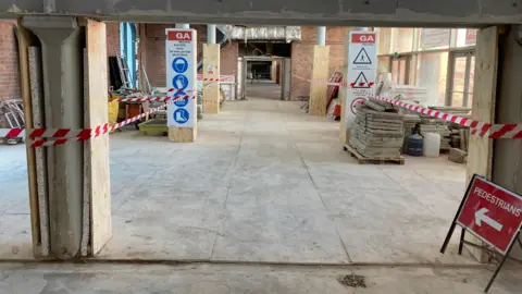 Refurbishment work taking place within the gallery. The space is sectioned off with red and white tape. There are piles of brick either side of a pedestrian walkway which is indicated by a red pedestrian sign.
