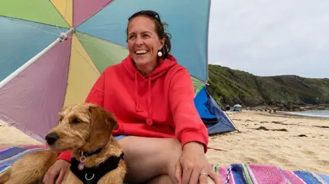 A woman wearing an orange hoodie is sat on a beach towel, on a beach, with her arm draped on a golden dog. They are sheltering under a beach umbrealla.