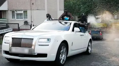 Three masked men are spraying Champagne from a white Rolls-Royce