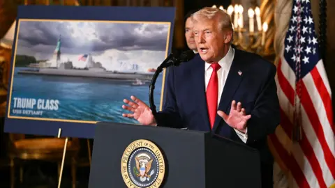 US President Donald Trump stood speaking at a lectern in a navy suit with red tie next to an image of a tanker