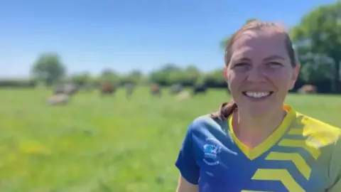 BBC A woman with tied back brown hair and a blue and yellow patterned sports top, standing in a field with the blurred shapes of animals in the distance