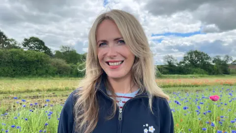 Evitts stood in a field of blue cornflowers, looking into the camera and smiling. She has long blonde hair, and is wearing a blue fleece with a white daisy logo embroidered on the chest. The sky is cloudy with some blue patches, and along the edge of the field are trees and bushes. 