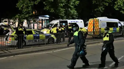 Getty Images Two police officers walking past the camera carrying helmets. In the background there are three police vans parked outside the Mercure Hotel at night, with around 10 police officers standing beside them