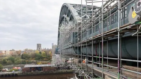 Newcastle City Council To the right, there is a large green metal arched bridge over the river, with scaffolding on it. In the distance there are trees and buildings and a car park. It is a cloudy day.