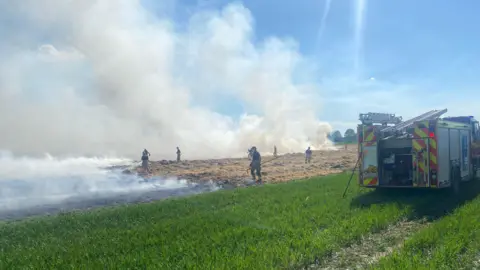 Firefighters tackling the fire in the field. A fire appliance can be seen to the right of the picture. There is a large amount of smoke. 