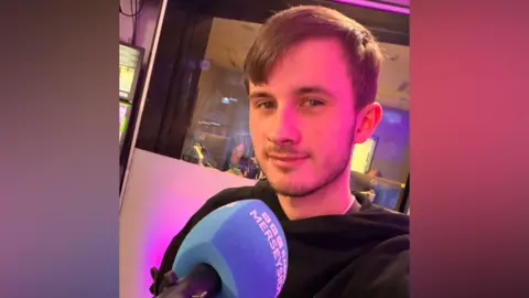 Luke Fitzpatrick has short brown hair and is wearing a black hooded top. He is inside the BBC Radio Merseyside studio and is sat behind a blue microphone