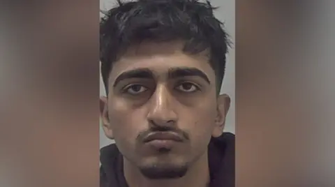 West Mercia Police A mugshot of a man with messy black hair and a moustache and goatee beard. He is wearing a black top.