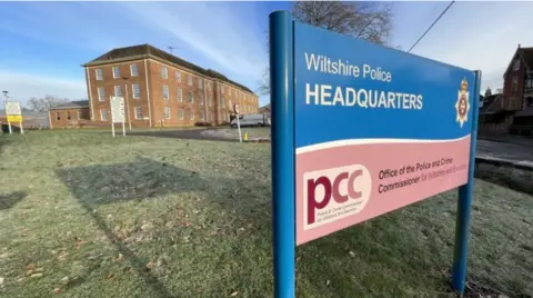 A sign which says 'Wiltshire Police headquarters' on grass with a building behind it