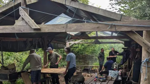 Six men are standing in a wooden building with open sides surrounded by trees. Three of them are studying a piece of wood on a worktable and the other three are talking to each other. On a wooden picnic bench in the foreground are metal plates, cutlery and drink cannisters.