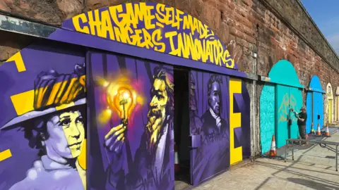 BBC One of the arches painted in yellow and purple, featuring a portrait of a woman and two men. At the top are the words 'Game-changers, self-made innovators". In the background, a man is spray painting the next arch along, which is painted in an aqua colour, with some golden graffiti.