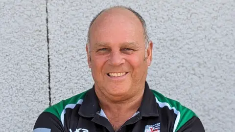 Headshot of Steve Worrall smiling, standing against a white outside wall, wearing a Chew Valley RFC top