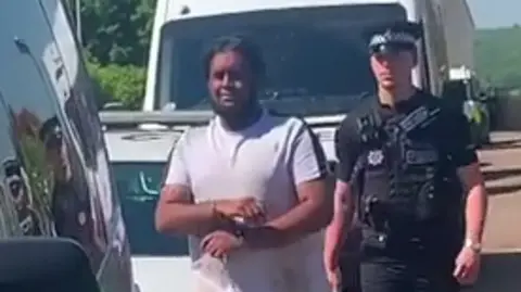 Supplied Oaveed Rahman is wearing a white T-shirt and is being marched to a police van by an officer in black uniform. 