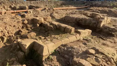 The image shows an archaeological excavation site with exposed stone foundations and partial walls made of weathered blocks. The ground is uneven and dusty, with trenches revealing rectangular room-like structures. Pieces of modern piping run across the top of the dig, contrasting with the historic remains below, and loose stones and rubble are scattered throughout the area.