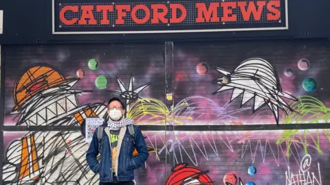 Robert Firth Zak Kilburn is wearing a mask, cap and denim jacket with a black and white scarf. He is stood facing the camera with the Catford Mews behind him. The shutters are covered in space-themed graffiti. 