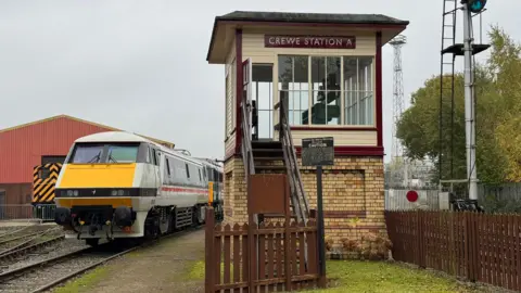 A railway museum - a train is visible on a track on the left and a small building for railway officers is on the right next to a brown-coloured fence. 