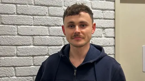 A man with short brown hair. He is looking directly at the camera and is wearing a blue zip up hoodie. 