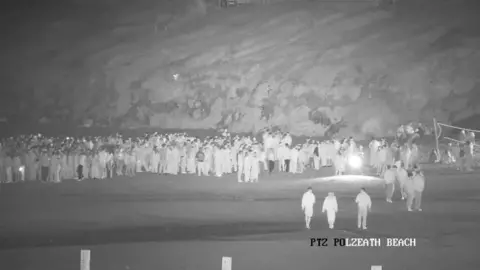A black and white image shows hundreds of young people gathered together on a beach. 