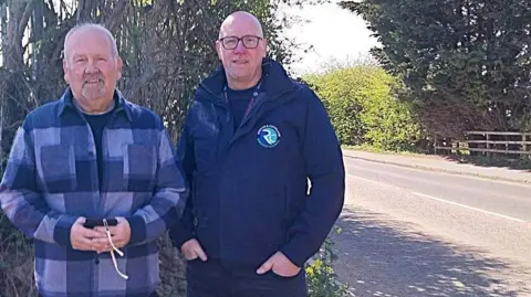 Councillor Stephen Crane and Councillor Carl Quartermain stood by Redcar Road. The stretch they are stood next to is lined by hedges and trees.