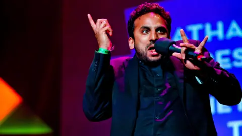 A man wearing all black speaking into a black microphone while making a dynamic pose; his right hand is pointing upwards while his left grips the microphone with his middle finger and his thumb. There are coloured stage lights in blue, orange, pink and green, lighting up the stage behind him and part on his suit.