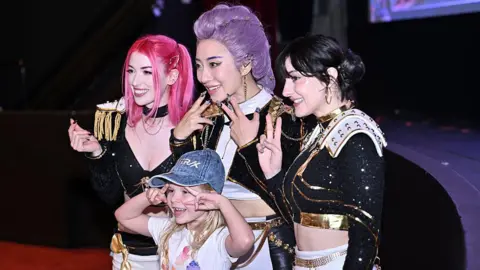 Getty Images Cosplayers dressed as Huntr/x from Kpop Demon Hunters pose for a photo with a young girl