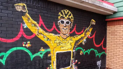 LDRS A colourful mural of Geraint Thomas wearing the yellow cycling jersey and helmet and white cycling glasses. Painted on a black background on a brick wall, with red and green swirls behind him.