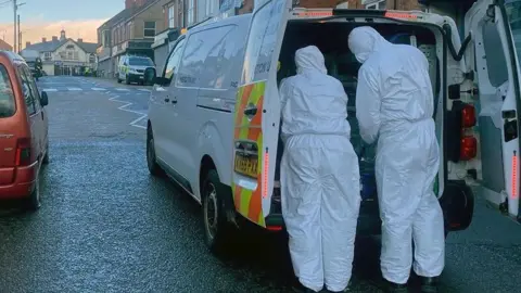 Two forensics officers dressed in white full-body suits are looking in the back of a police van. Another van can be seen further up the street.