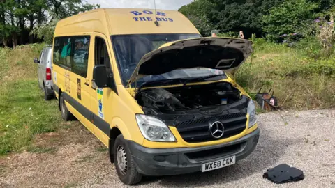 Yellow minibus with its bonnet open, parked on a gravel driveway beside trees and greenery.
