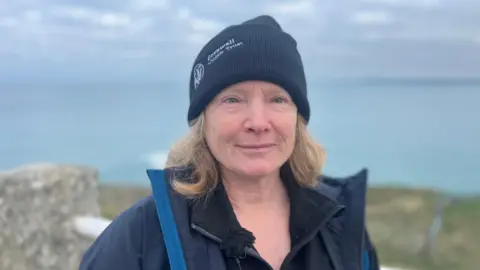 A close-up picture of Bex Allen in front of the coastline. She has blonde hair and is wearing a black beanie hat which says 'Cornwall Wildlife Trust' on it. She is smiling at the camera.