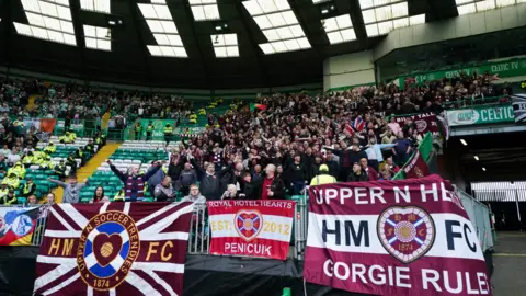 PA Media Hearts supporters sing and wave flags inside Celtic Park, with large maroon banners hanging over the front railing as home fans fill the stands behind them.