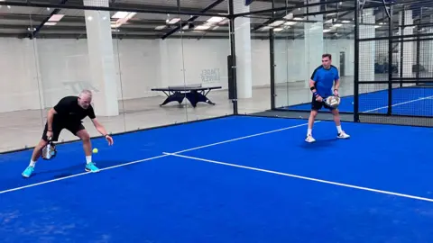Sam Read/BBC Two men during a game of padel at an indoor centre. The back wall of the court is a glass panel and the court itself is blue with white markings.
