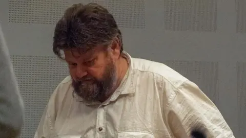 Carl Beech, a man in his 50s with dark grey hair and a mid-length beard, walking along with his head down, wearing a cream-coloured shirt.