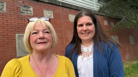 Two women stand side by side in front of a red brick wall. The one of the left has blonde hair, has reading glasses pushed onto her head and is wearing a mustard top. On the right the woman wears a blue cardigan over a white top which separates into a floral pattern around the neck. 