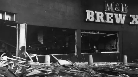 Getty Images A black and white image of one of the pubs that was blown up in the city. Debris is on the floor with windows blown out below a M&B sign.