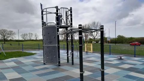 North Tyneside Council Blue and grey checkboard type rubber paving under a large climbing frame. There's blue fencing around it and grass in the background.