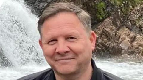 A man smiling at the camera. He is standing in front of a waterfall. He is wearing a blu et-shirt and a black casual jacket. 
