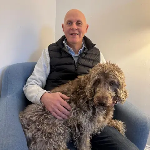 Gary Tait sits in a light blue fabric chair against a plain cream wall. He is wearing a blue shirt, grey jumper and a padded gilet. A big shaggy light brown dog sits on his knee and Gary has a hand across its curly fur.