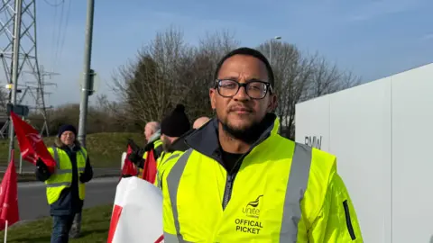 A man with short hair and a beard stands on the picket line outside the BMW Hams Hall plant in Coleshill. He is wearing glasses and a high-visibility, yellow jacket with the words Unite the Union Official Picket