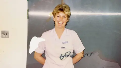 Sandra Bourgaize An old photo of Sandra Bourgaize which shows a women with short blonde hair in a white or light pink nurses scrubs stood in front of silver metal doors. There is some markings on the photo from wear and tear.