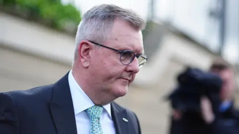 A side on image of a man with short grey hair. He is wearing black and blue rimmed round glasses. He is wearing a dark suit, white shirt and turquoise patterned tie. He has a badge in the shape of a fish pinned to his jacket. The background is blurred.