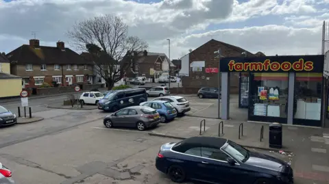 George Carden/BBC Cars parked in the car park which is in a suburban area, one shop FarmFoods is visible