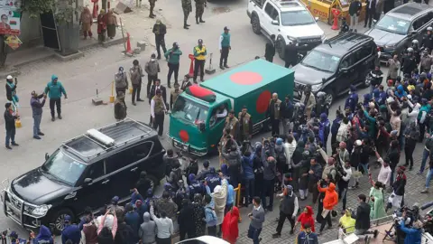 AFP via Getty Images Security personnel escort a motorcade carrying Bangladesh's former prime minister Khaleda Zia's remains from Evercare hospital in Dhaka. People are seen reaching out with the phones to take photographs of the hearse draped with the Bangladesh national flag