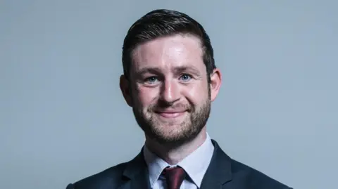 A man with short, dark brown hair and facial hair smiling. He is wearing a dark blue suit jacket, light blue shirt and a red tie, stood in front of a grey background.