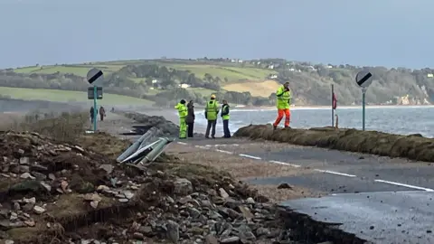 The A379 Slapton Line between Torcross and Slapton has washed away. People in high-visibility clothing are seen on the road.