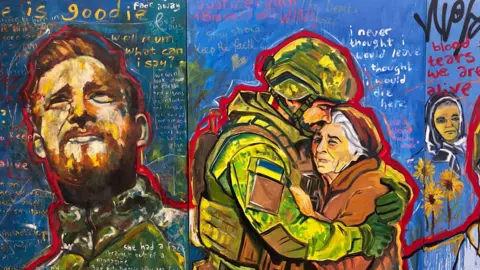 A painting showing Ukrainian people affected by the war. In one scene, a Ukrainian soldier looks into the distance and, in another, a different Ukrainian soldier hugs an old woman wearing an orange coat and hat. The background is blue and there is emotive writing surrounding the figures.