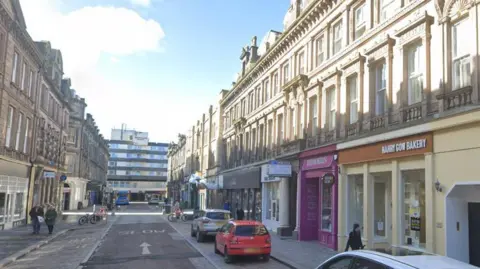 A street with historic facades and colourful shop fronts on a sunny day with cars parked on both sides of the road