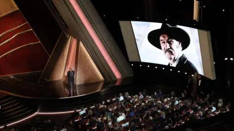 Getty Images Morgan speaks at the Oscars as a shot of Hackman is projected onto the screen. An audience watches