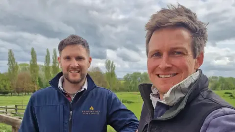 The image shows two people standing outdoors by a wooden fence in a green countryside setting. Both individuals are wearing casual outdoor clothing, including jackets or vests. One person is holding the camera for a selfie while the other leans on the fence. Behind them are grassy fields, scattered trees, and a cloudy sky. Visible text on one jacket reads “PANPWNTON FARM CAMPSITE.”